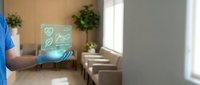 Male healthcare professional in blue scrubs holding digital tablet displaying holographic medical icons and heart symbols in modern clinic interior with plants and natural lighting.