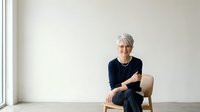 Senior woman with gray hair wearing dark navy cardigan, sitting on wooden chair against white wall background, isolated on white for design use.