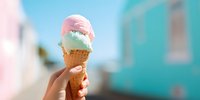 Hand holding a colorful ice cream cone with pink and mint scoops against a blurred turquoise building background on a sunny summer day.