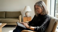 Mature Caucasian woman with gray hair reading tablet computer while relaxing in comfortable armchair at home during daytime.