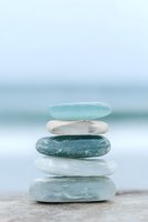 Stacked smooth stones in shades of turquoise, white, and gray balanced on sandy beach with blurred ocean backdrop, symbolizing zen, balance, and tranquility for wellness and meditation designs.