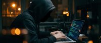 Hooded hacker typing on laptop computer in dark room with glowing city lights. Cybersecurity threat concept for digital crime and data breach awareness campaigns.