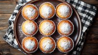 Freshly baked vanilla muffins dusted with powdered sugar on rustic wooden plate with checkered napkin, homemade pastry photographed from above.