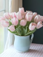 Fresh pink tulips arranged in vintage white ceramic pitcher on checkered tablecloth near window creating elegant spring home decor and floral arrangement.