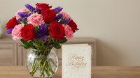 Colorful mixed flower bouquet with red roses, pink carnations and purple statice in glass vase next to Happy Birthday greeting card on wooden table.
