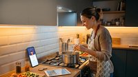 Woman in casual home attire cooking on stovetop while viewing recipe on smartphone, modern kitchen with warm lighting and subway tile backsplash for culinary content.