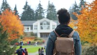 Young Asian male student with backpack looking at college campus building surrounded by autumn trees and fellow students on lawn. Fall education concept.