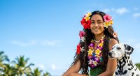 Young woman with long dark hair and South Asian ethnicity wearing vibrant floral lei and flower crown, holding spotted white dog, tropical setting with palm trees and blue sky.