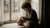Young boy with light brown hair, Caucasian ethnicity, sitting by window playing with piggy bank, learning about saving money and financial literacy in home setting.