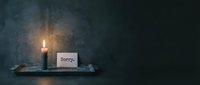 Lit dark candle on a tray beside a sorry note card against a moody gray concrete wall, conveying apology, regret, and sympathy.
