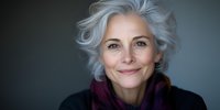 Confident mature Caucasian woman with silver gray hair and natural smile wearing dark sweater and scarf against blurred background, representing aging gracefully.