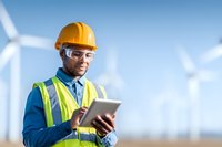 African American male engineer in safety vest and hard hat using digital tablet at wind turbine farm, representing renewable energy and sustainable development.