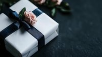 White gift box wrapped with navy blue ribbon and decorated with fresh pink rose on dark stone background, elegant present packaging for special occasion.