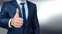 Businessman in navy blue suit showing thumbs up gesture, conveying approval and positive feedback in professional setting against gradient background.