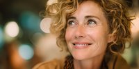 Mature Caucasian woman with curly blonde hair smiling and looking up with hopeful expression against blurred bokeh background.