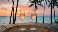 Two champagne glasses on tropical beach table at sunset with palm trees and ocean view for romantic celebration and vacation dining.