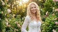 Blonde bride in white lace wedding dress smiling in blooming rose garden with soft sunlight filtering through greenery for romantic wedding photography.