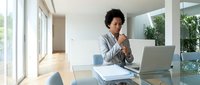 Young African American businesswoman in gray suit working on laptop at modern office desk, professional corporate environment with natural lighting.
