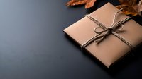 Gift box wrapped in kraft paper with rustic twine bow and autumn dry leaves on dark background, minimalist holiday present packaging composition.