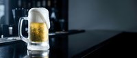 Cold beer mug with golden lager and foam head on dark bar counter in modern pub setting for hospitality and beverage marketing materials.