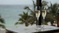 Two champagne flutes filled with sparkling wine on marble table with tropical ocean view and palm trees in blurred background for luxury vacation celebration.