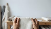 Hands holding natural linen fabric swatch with wooden rack and rolled textiles in background, suitable for textile design, interior styling, and craft projects.