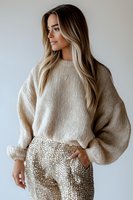 Young Caucasian woman with long blonde balayage hair wearing oversized beige knit sweater and gold sequin pants poses against white wall in modern fashion portrait.