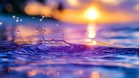 Water drop splash creating circular ripples on blue water surface against golden sunset sky, with bokeh light effects and reflections on calm ocean waves.
