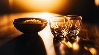 Atmospheric still life with black ceramic bowl and crystal glasses on wooden table illuminated by warm golden sunset light through window. Moody dining setup.