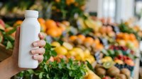 Close-up of hand holding blank white bottle at farmers market with fresh produce display in blurred background for beverage packaging mockup.