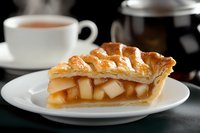 Fresh baked apple pie slice with flaky golden crust and caramel sauce on white plate, served with hot tea against dark background. Perfect for food blogs.