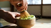 Young adult with medium skin tone using traditional bamboo whisk to prepare matcha tea powder in wooden bowl with fresh green matcha and water, traditional Japanese tea ceremony preparation for culinary, wellness and cultural content.