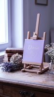 Wooden easel with lavender canvas displaying dream big script, surrounded by dried flowers and vintage books on a rustic wooden desk.