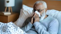 Senior African American man with gray beard blowing nose while lying sick in bed at home, wearing light blue sweater, experiencing cold or flu symptoms.