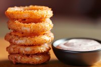 Crispy golden onion rings stacked in tower formation with creamy dipping sauce in black bowl. Close-up shot with selective focus and warm lighting.