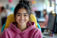 Young Asian woman wearing pink hoodie smiling at camera in modern office environment. Natural daylight illuminates cheerful expression and casual workplace setting.