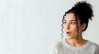 Young woman with dark curly hair styled in a bun, medium skin tone, wearing a light gray ribbed sweater, looking to the side with a surprised or thoughtful expression against a white background.