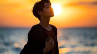 Young Asian man in black clothing standing by ocean at golden sunset, eyes closed in peaceful meditation pose against dramatic orange sky and sparkling water background.