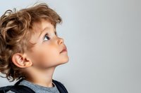 Young Caucasian boy with curly brown hair looking up with wonder and curiosity against light gray background, wearing casual denim clothing, profile view portrait.