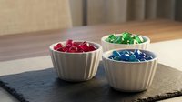 Three white ceramic bowls filled with colorful red, green, and blue candies arranged on a dark slate board on a wooden table.