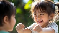 Happy Asian toddler girl laughing and playing outdoors with friend in sunny garden setting. Natural childhood joy and friendship moment for family content.