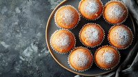 Fresh homemade vanilla muffins dusted with powdered sugar served on dark plate against textured gray background with linen napkin, top view.