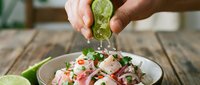 Hands squeezing fresh lime juice over ceviche with seafood, vegetables and herbs in white bowl on wooden table for cooking and food preparation.
