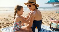 Mother applying sunscreen to daughter on beach during summer vacation. Woman in hat and sunglasses protecting child from UV rays on sandy shore with ocean waves.