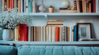 Books arranged on white wooden shelf with teal vase holding baby's breath flowers, creating cozy home library atmosphere against soft teal wall background.
