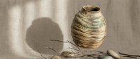 Rustic handmade ceramic vase with textured surface beside dry twigs and pebbles on linen background with natural shadows.