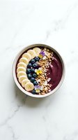 Acai smoothie bowl topped with banana slices, blueberries, granola, and edible flowers on a marble surface, overhead view.