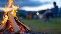 Burning campfire with wooden logs at dusk, glowing flames and embers with blurred people relaxing in background at camping site. Outdoor adventure atmosphere.