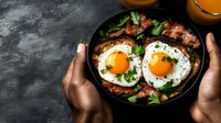 Sunny side up eggs with crispy bacon in cast iron skillet held by dark-skinned hands against black background, fresh herbs and orange juice for breakfast.