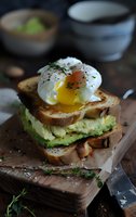 Poached egg with runny yolk on toasted bread and mashed avocado, seasoned with herbs and black pepper, served on rustic wooden board. Food photography.